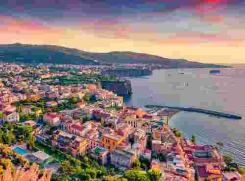 Aerial view of a colorful coastal town near Naples, with pastel houses clustered along cliffs above the sea and the coastline stretching into the distance at sunset.