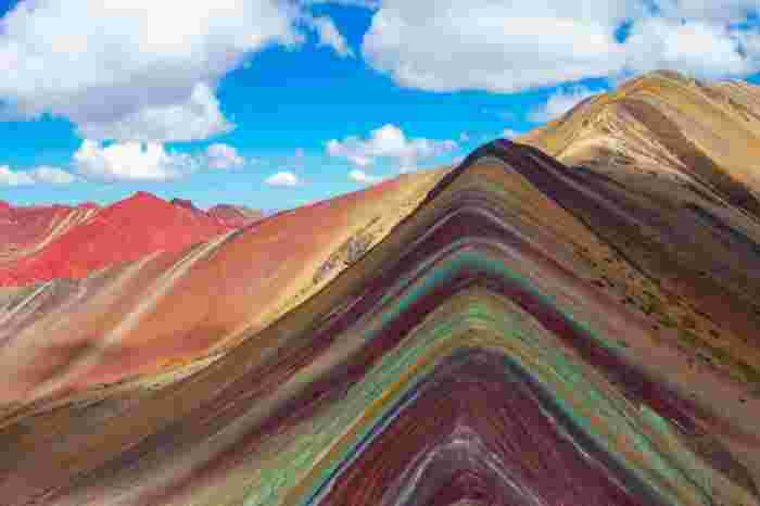Colorful layered slopes of Vinicunca (Rainbow Mountain) in Peru under a bright blue sky with scattered clouds.