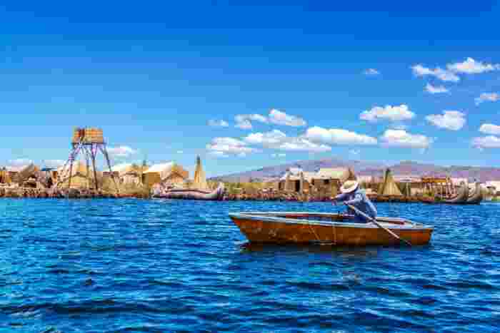 Person rowing a small wooden boat on Lake Titicaca with traditional reed houses and structures in the background.