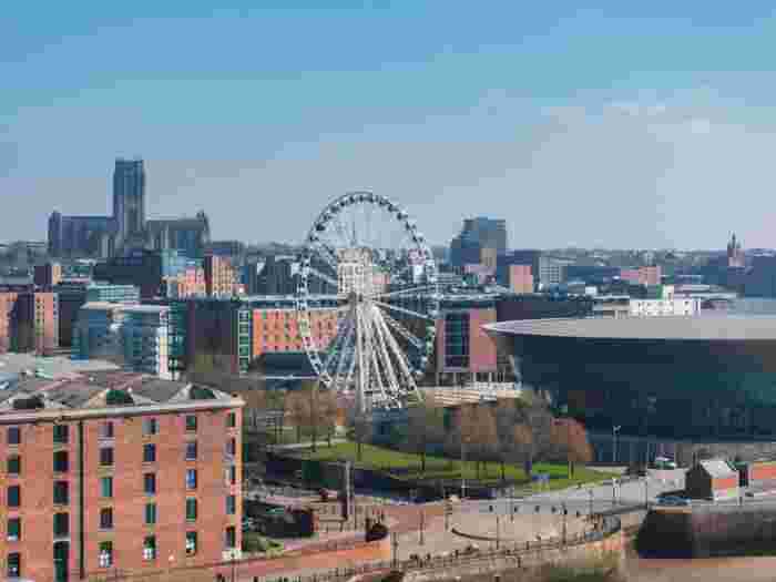 image Liverpool waterfront skyline featuring a large Ferris wheel, the Echo Arena, and Liverpool Cathedral under a clear blue sky.