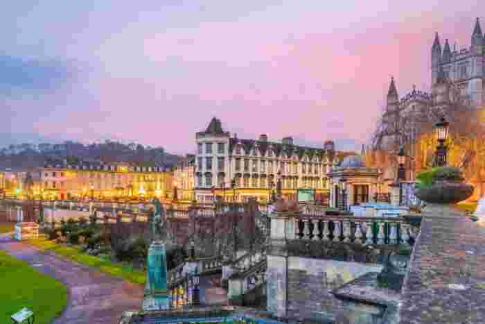 image Bath city center at sunset with Georgian architecture, glowing street lamps, and Bath Abbey's Gothic spires rising against a pink sky.