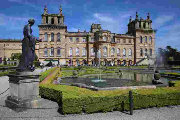 image Blenheim Palace's grand Baroque facade with ornamental fountains, manicured hedges, and a bronze statue in the foreground.