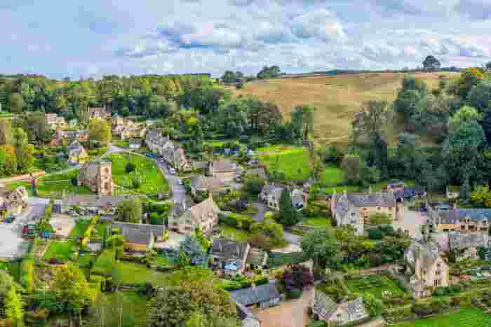 image Aerial view of a picturesque Cotswolds village with honey-stone cottages, a small church, and lush green hills in the English countryside.