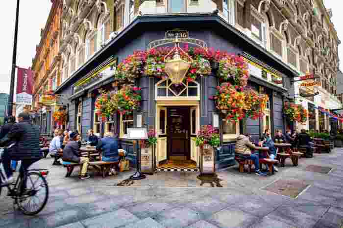 image A charming traditional English pub on a London street corner decorated with colorful hanging flower baskets, with patrons seated outside.