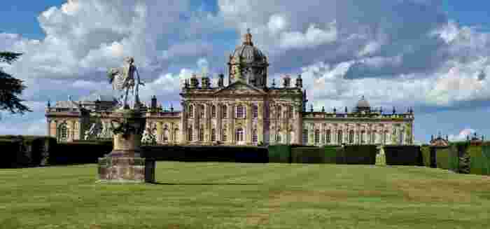 image Castle Howard's Baroque facade with a central dome, surrounded by manicured hedges and a classical stone statue on the lawn.