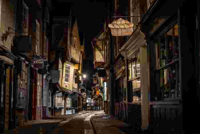 image The Shambles in York at night, a narrow medieval lane with overhanging buildings and warm lamplight casting shadows on the empty cobblestones.