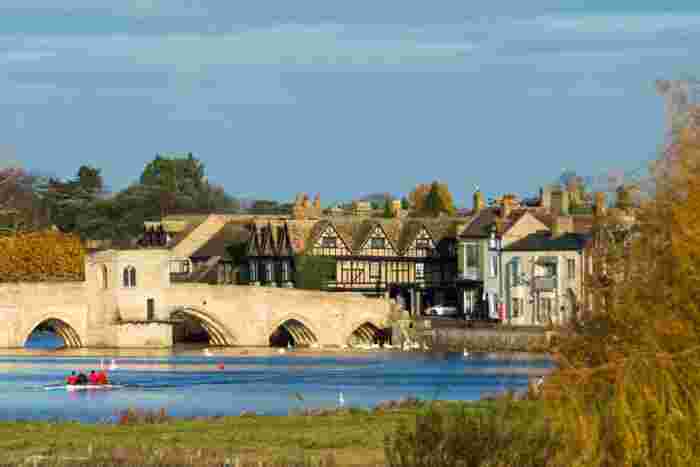 image A stone arched bridge over a river with a rowing team on the water, swans nearby, and traditional English buildings along the bank in autumn.