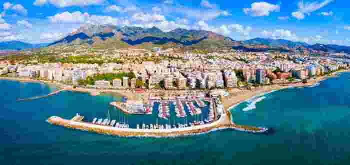 Aerial panorama of Marbella’s coastline, marina, sandy beaches, and mountain backdrop on a sunny day. 
