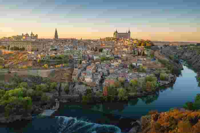 Scenic panorama of Toledo at sunset with the Tagus River, medieval skyline, and Alcázar rising above the city. 