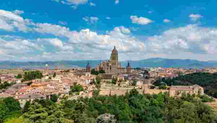 Panoramic view of Segovia with its historic old town, cathedral, and surrounding green hills under a bright sky. 