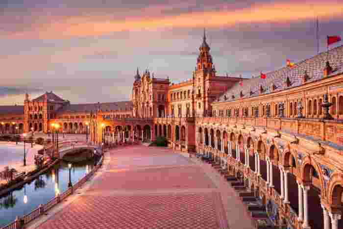 Plaza de España in Seville at dusk with ornate arches, tiled bridges, and reflections along the canal. 