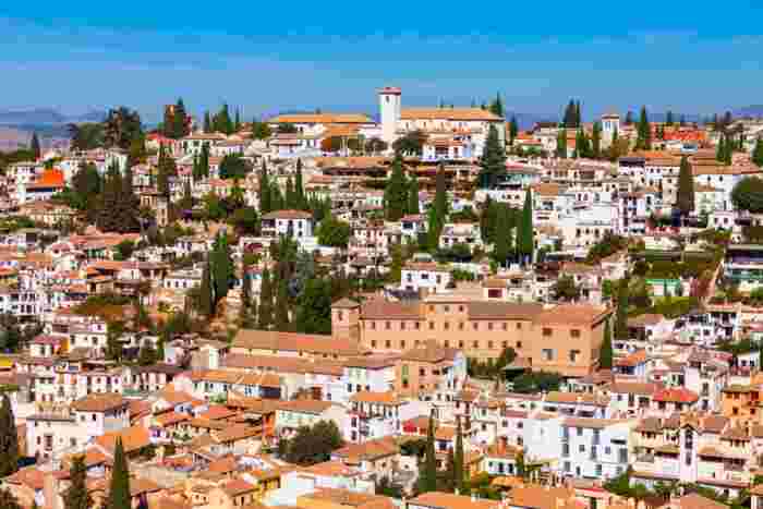 Panoramic view of Granada’s Albaicín neighborhood with whitewashed houses, cypress trees, and rolling hills under a clear blue sky. 