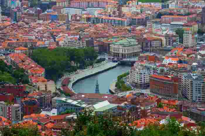 Panoramic cityscape of Bilbao with the Nervión River, red rooftops, and urban buildings stretching across the valley. 