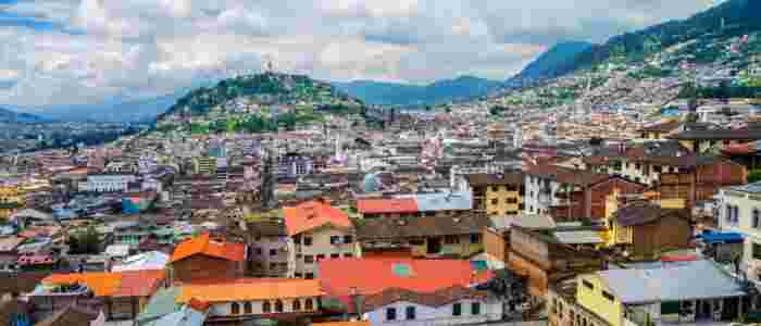 Panoramic aerial view of Quito, Ecuador, with colorful hillside neighborhoods, colonial architecture, and the Andes Mountains under cloudy skies 