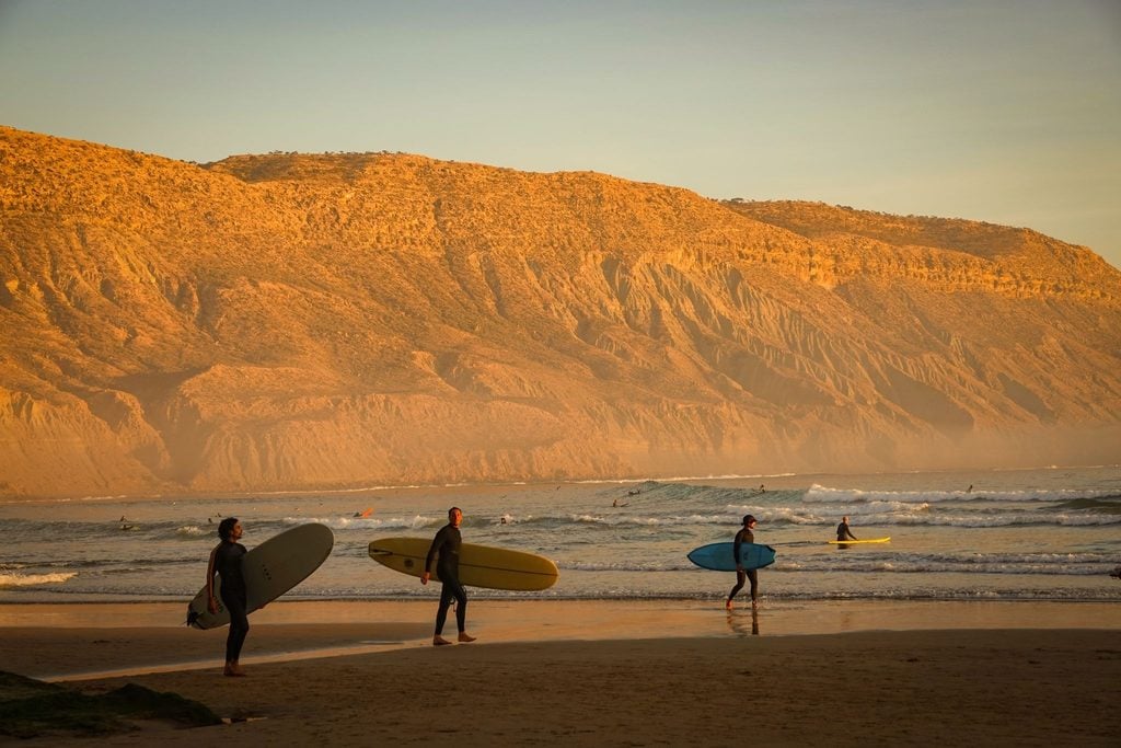 Surfers carrying boards along a golden beach beneath sunlit cliffs at dusk 