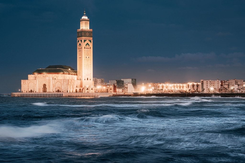 Illuminated Hassan II Mosque tower beside a stormy sea at dusk 