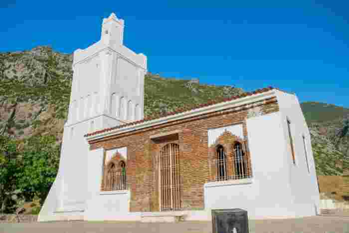 Whitewashed mosque with a tall square minaret and brick-arched windows, set against a rocky mountain backdrop.