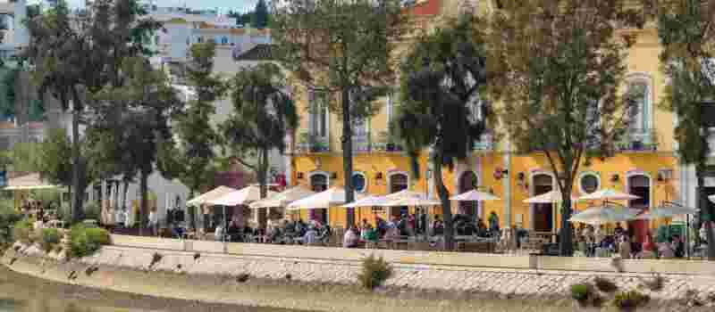 Outdoor riverside café terrace with white umbrellas in front of a yellow Baroque-style building along the Gilão River in Tavira, Algarve
