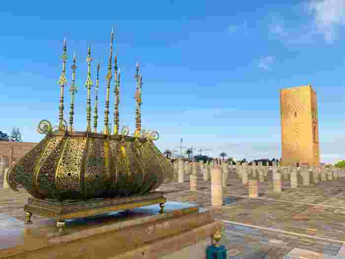 An ornate gilded brass decoration on the Mohammed V Mausoleum esplanade, with the Hassan Tower and its column field behind.