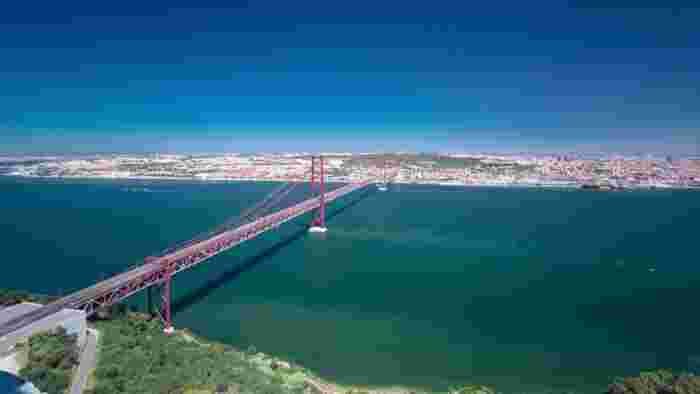 The red 25 de Abril Bridge crossing the wide Tagus River, with the Lisbon skyline on the far bank under a deep blue sky