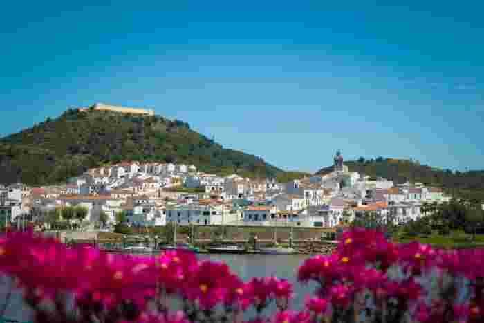 Whitewashed hillside village of Sanlúcar de Guadiana with a fort on the hilltop, seen across the Guadiana River with moored sailboats and pink flowers in the foreground