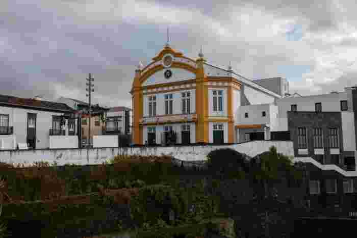 Yellow and white theater building with an ornate gabled facade above a dark volcanic stone wall under a cloudy sky in the Azores