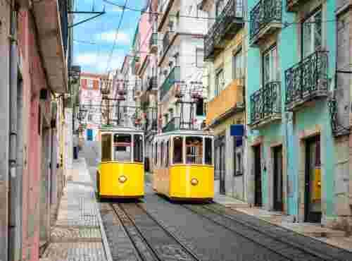 Two yellow Bica funicular cars passing each other on a steep cobblestone street lined with colorful pink, yellow and turquoise buildings with wrought-iron balconies in Lisbon