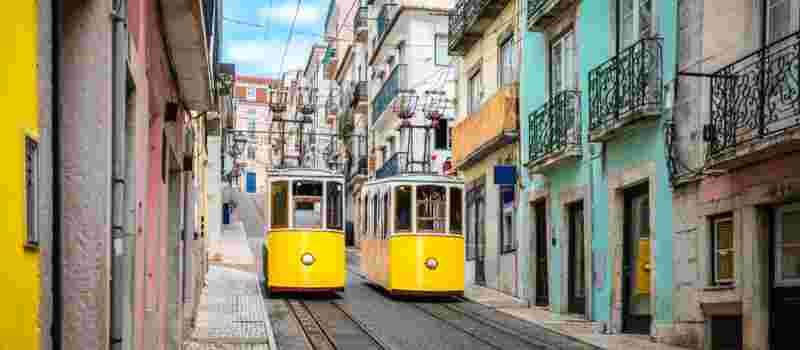 Two yellow Bica funicular cars passing each other on a steep cobblestone street lined with colorful pink, yellow and turquoise buildings with wrought-iron balconies in Lisbon