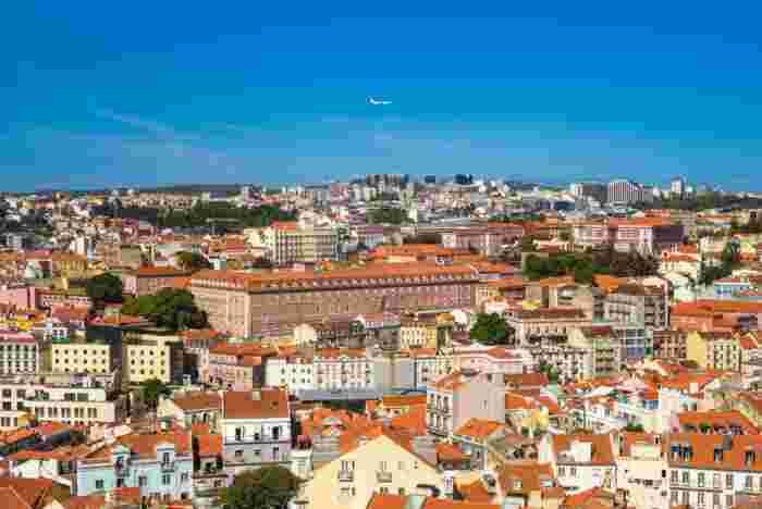 Aerial view of Lisbon's terracotta rooftops and pastel-colored buildings, with an airplane flying across the blue sky