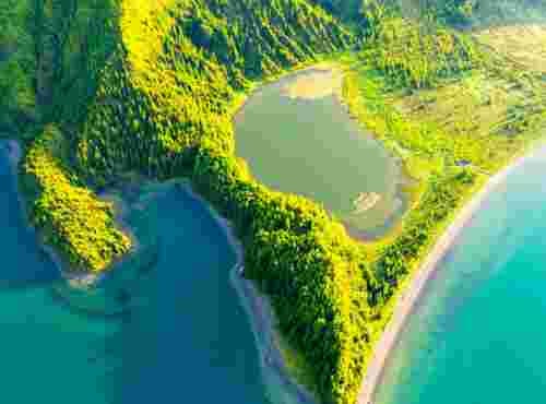 Aerial view of a green forested volcanic ridge on São Miguel in the Azores, with a small lagoon beside a curved gray sand beach and turquoise crater lake water on both sides