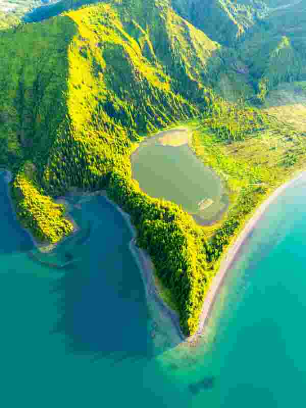 Aerial view of a green forested volcanic ridge on São Miguel in the Azores, with a small lagoon beside a curved gray sand beach and turquoise crater lake water on both sides