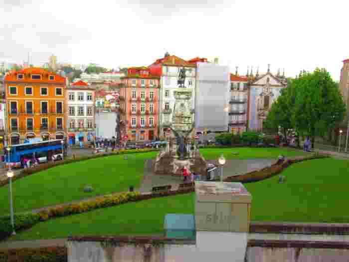 Rainy day in a Porto square with a bronze monument on a green lawn, colorful buildings, a double-decker bus and people with umbrellas, with the Douro River in the background