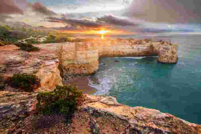 Natural rock arch over a turquoise cove at Arco de Albandeira in the Algarve, with the sun low on the horizon above the sea