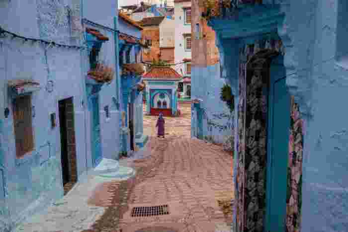 Narrow blue-washed alley in Chefchaouen leading to a decorative fountain, with a woman in a purple robe walking toward it.