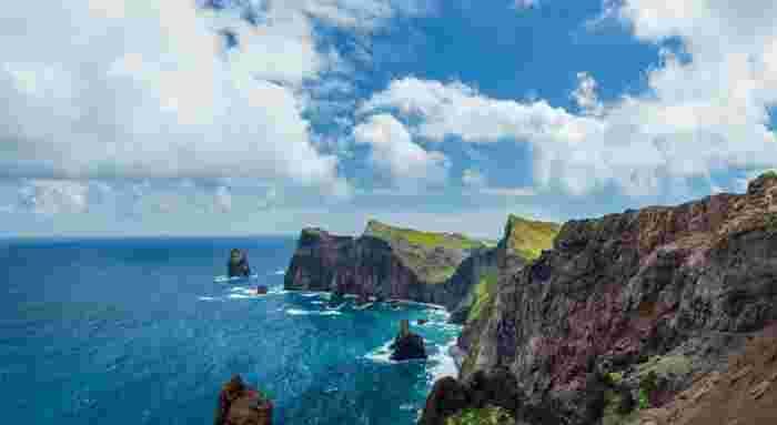 Rugged red and brown sea cliffs with green tops and rock stacks rising from the deep blue Atlantic Ocean at Ponta do Rosto in Madeira