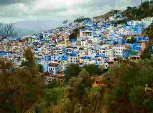 Aerial view of Chefchaouen's blue-and-white hillside houses spread across a mountainside beneath cloudy skies.