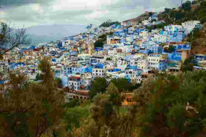 Aerial view of Chefchaouen's blue-and-white hillside houses spread across a mountainside beneath cloudy skies.