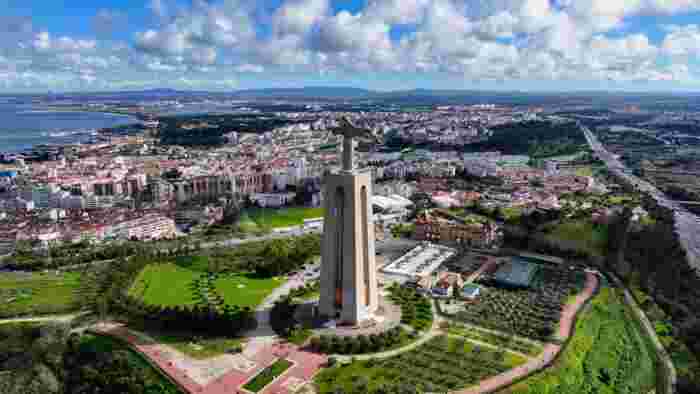 Aerial view of the Christ the King statue on its tall pedestal in Almada, surrounded by gardens, with the rooftops of the city and the Tagus River behind